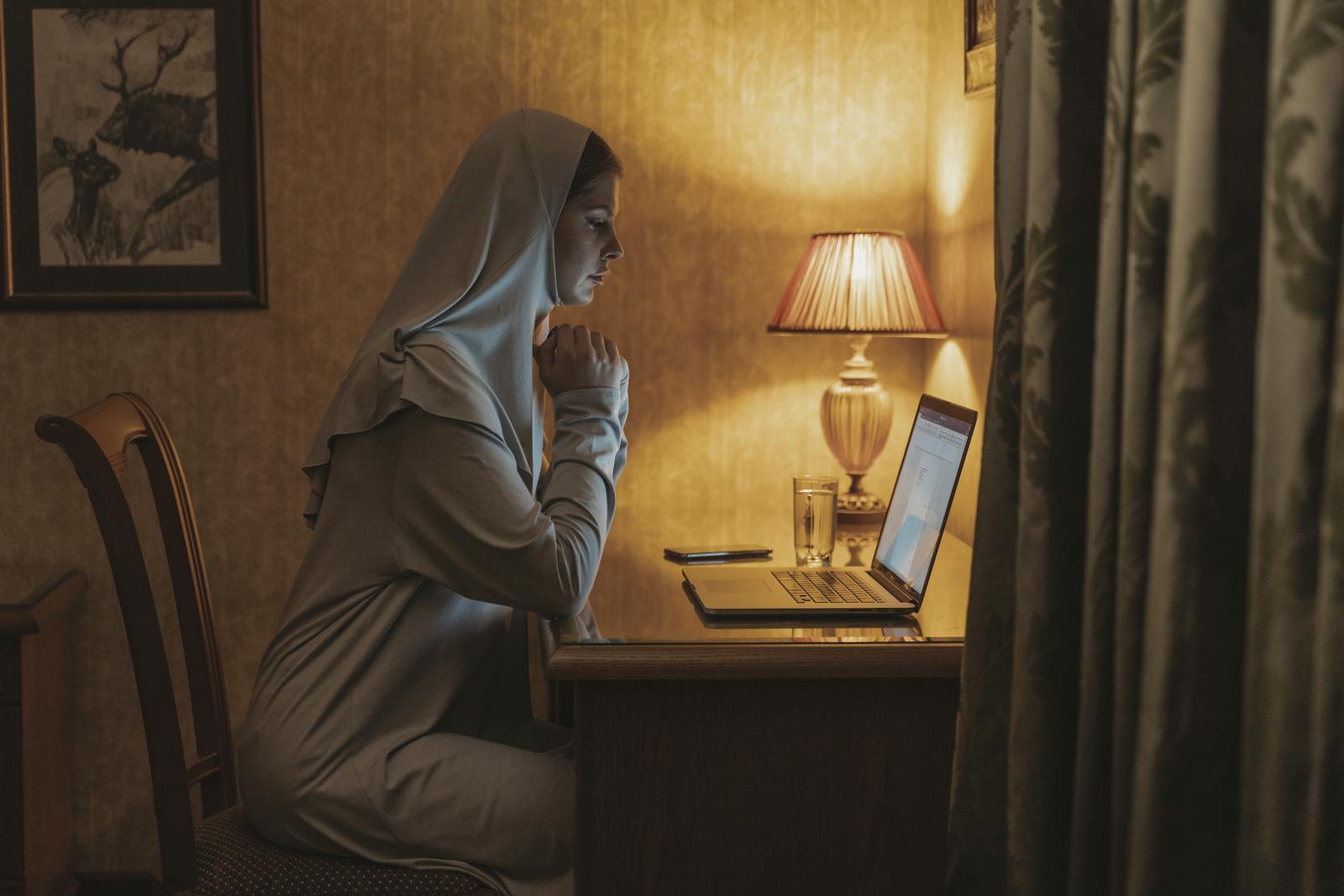 Woman in hijab working remotely at home on laptop during nighttime.